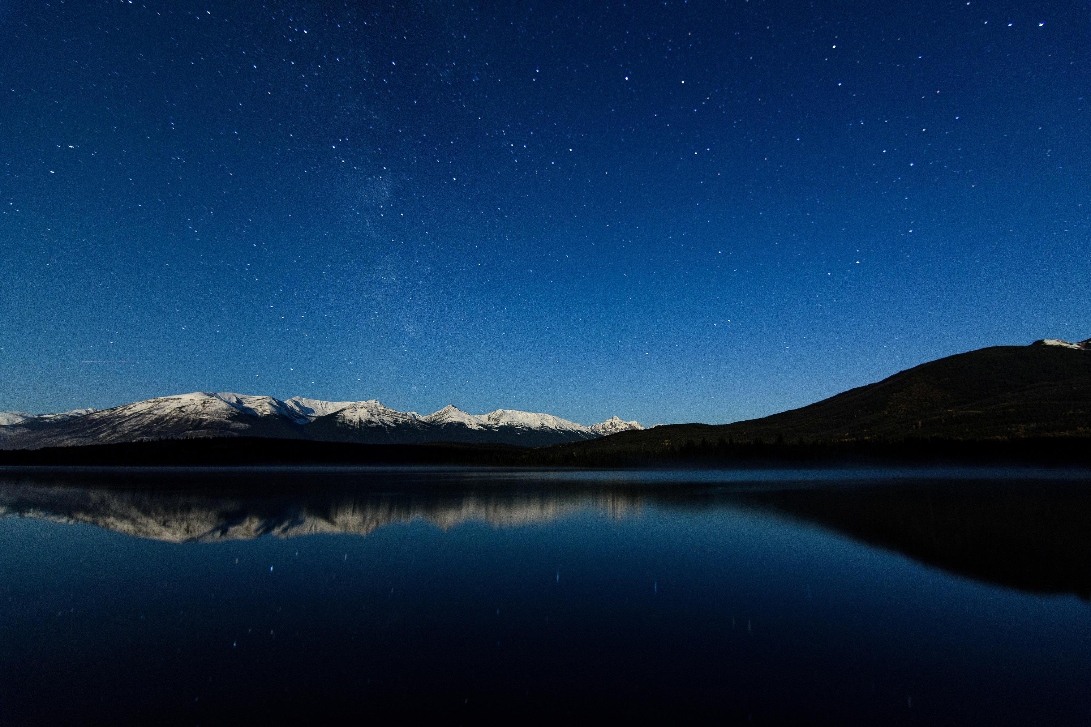 The blue, starry night sky, striped by the vertical rise of the Milky Way, rises above a snow-capped mountain range, sloping gently up from left, to a range, then back down again just right of centre and back up again to a taller mountain. Below, a still, deep lake reflects back the sky and mountains and flanking forest as in a mirror. The blue, starry night sky, striped by the vertical rise of the Milky Way, rises above a snow-capped mountain range, sloping gently up from left, to a range, then back down again just right of centre and back up again to a taller mountain. Below, a still, deep lake reflects back the sky and mountains and flanking forest as in a mirror.